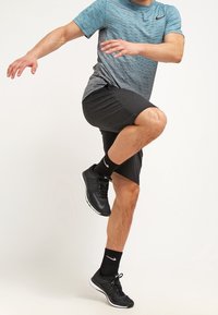 Conjunto deportivo que presenta una camiseta de manga corta azul y gris, pantalones cortos negros, zapatillas negras y calcetines negros, mostrando una pose activa con la rodilla levantada.