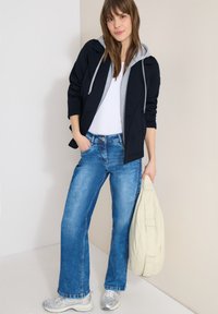 Femme portant une veste noire avec une capuche grise, une chemise blanche, un jean bleu et des baskets argentées, tenant un sac matelassé blanc à côté d'elle.