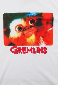 Valkoinen t-paita, jossa on värikäs grafiikka suurella silmäisellä olennolla, jolla on ilkikurinen ilme, sekä teksti "GREMLINS" rohkealla punaisella alla.