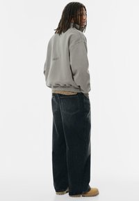 Šedá fleecová bunda s vysokým límcem, volné tmavé džíny a světle hnědé boty. Bunda má na zadní straně logo. Jednoduchý, uvolněný design.