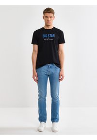 Hombre joven de pie, vistiendo una camiseta negra con la inscripción "BIG STAR", jeans azul claro y zapatillas blancas, sobre un fondo blanco liso.