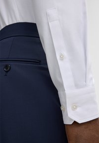 Close-up van de arm van een persoon in een witte, gestructureerde shirtmanchet met knopen, naast marineblauwe op maat gemaakte broek met een achterzak met knopen.