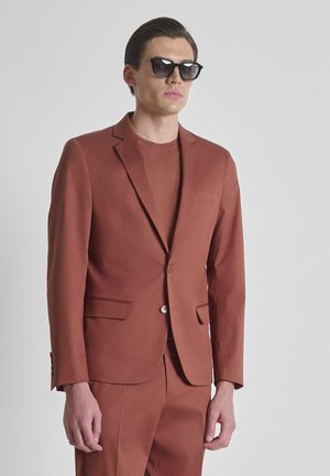 Mannelijk model draagt een roestkleurig pak en trui met zwarte zonnebril, staand tegen een effen lichtgrijze achtergrond.