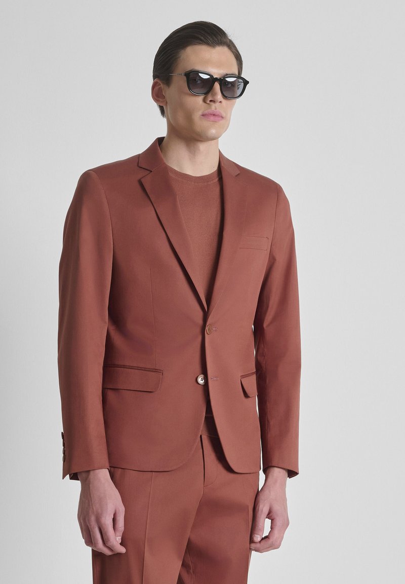 Modello maschile che indossa un abito color ruggine e un maglione, con occhiali da sole neri, in piedi davanti a uno sfondo grigio chiaro uniforme.