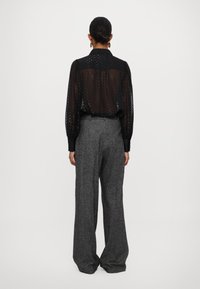 Zwarte, transparante blouse met lange mouwen en een gestippeld patroon, gecombineerd met hoge, wijde grijze broek van textuurstof.