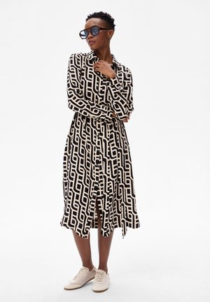 Kobieta w czarno-białej sukience midi w geometryczny wzór, w sneakersach i dużych okularach przeciwsłonecznych, stojąca z założonymi ramionami.