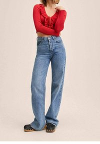 Maglietta rossa a maniche lunghe con collo arricciato; jeans a vita alta azzurri chiari a gamba larga; zoccoli neri con suole in legno e dettagli argentati.