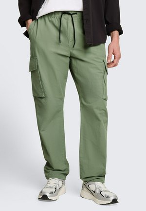 Mężczyzna w jasnizołych spodniach cargo z czarnym sznurkiem, białych tenisówkach, czarnej kurtce i białej koszuli, stoi z rękami w kieszeniach.