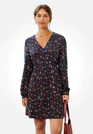 Robe fleurie bleu marine à manches longues, encolure en V, détail de boutons et motifs floraux multicolores. Assortie avec un sac à main rouge.