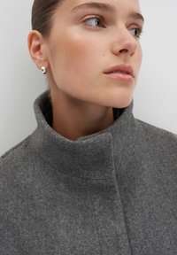 Grauem Wollmantel mit hohem Kragen und klarer Linienführung. Das Model trägt einen runden silbernen Ohrring; der neutrale Hintergrund hebt die Produkteigenschaften hervor.