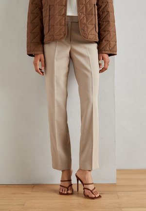 Mujer con pantalones beige de pierna recta, chaqueta marrón acolchada y sandalias marrones de tacón alto con tiras, de pie sobre un suelo de madera.