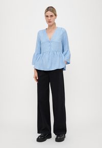 Svetlo modra peplum majica z V-izrezom, gumbom spredaj in razširjenimi rokavi; teksturirano blago. V par z črnimi širokimi hlačami in črnimi masivnimi čevlji.