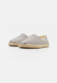 Des chaussures slip-on en toile grise avec une semelle en jute, présentant une texture lisse et un détail de couture en croix sur le dessus.
