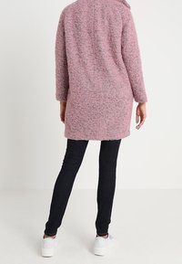 Personne portant un manteau texturé rose à manches longues, un pantalon noir ajusté et des baskets blanches, debout de dos face au spectateur.