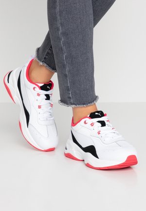 Weiße Turnschuhe mit schwarzen und roten Akzenten, getragen zu grauen Jeans mit ausgefranstem Saum, auf einer schlichten weißen Oberfläche.