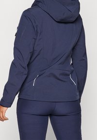 Veste imperméable bleu marine avec capuche, dotée d'un design sans coutures, de fermetures éclair blanches et d'une poche latérale sur le bras gauche.
