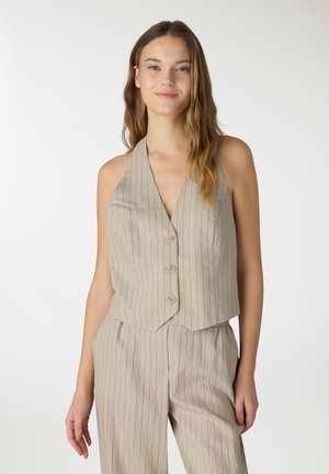 Donna con capelli lunghi che indossa un gilet beige a righe sottili senza maniche con bottoni e pantaloni coordinati a vita alta, in piedi davanti a uno sfondo semplice.