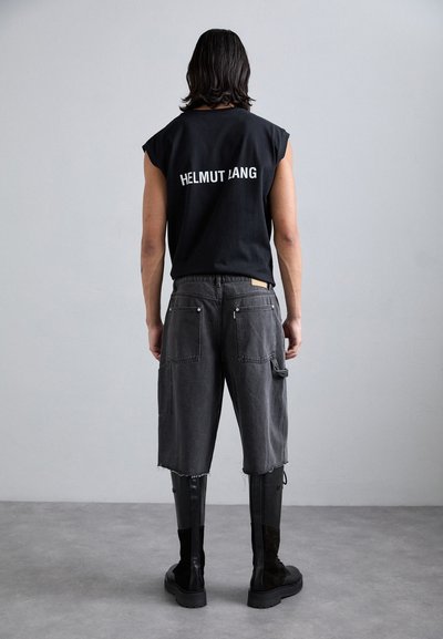 Zwarte mouwloze shirt met het logo "HELMUT LANG", grijze cargoshorts en zwarte kniehoge laarzen met gestructureerde zolen, met de rug naar de camera.