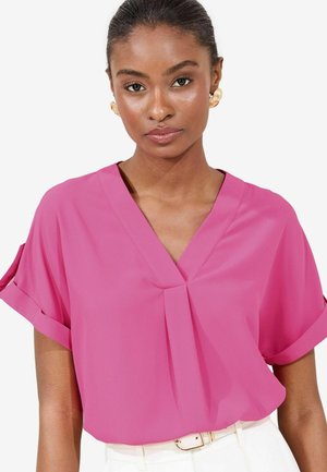 Camicetta rosa a maniche corte con scollo a V e dettaglio plissettato sul davanti. Realizzata in tessuto leggero. Abbinata a una cintura crema.