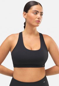 Sujetador deportivo negro con escote en forma de barco y tirantes anchos. La tela parece suave y elástica, diseñada para ropa deportiva. Detalle sutil del logotipo en el dobladillo.