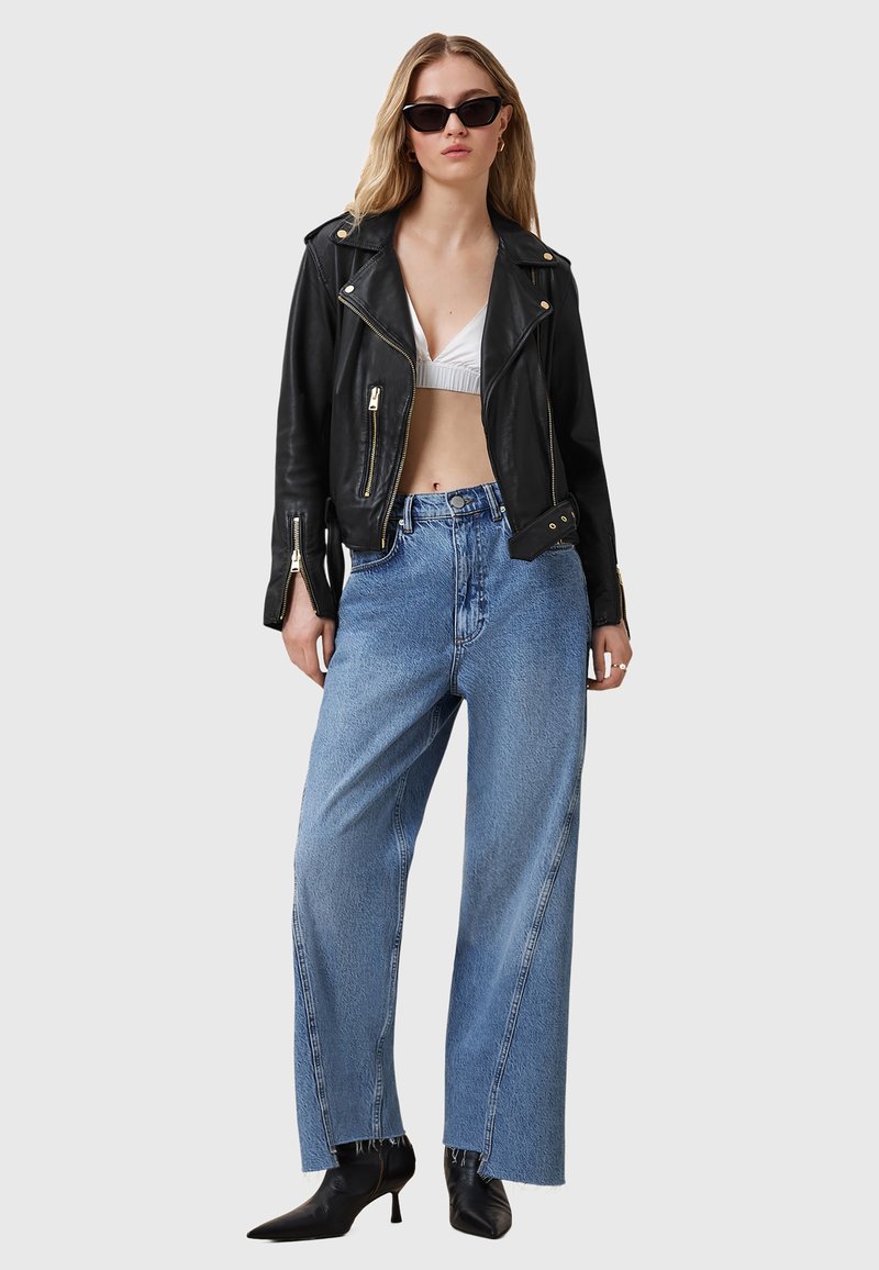 Giacca da motociclista nera in pelle, bralette bianca, jeans in denim azzurro a gamba larga e stivaletti neri a punta con finitura lucida. Occhiali da sole neri.