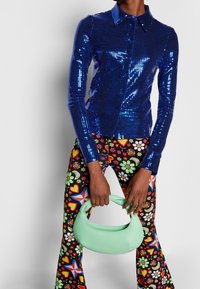 Camisa azul con lentejuelas y cierre de botones, combinada con pantalones de campana negros con estampado floral y un bolso redondeado en verde menta.