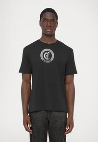 Mees, kes kannab musta T-särki, millel on valge ringshape madu logo ja "Just Cavalli" tekst, seistes ühtlase valge tausta ees.