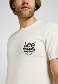 Camiseta de algodón color crema de manga corta, con un logo negro que dice "Lee Jeans Quality Denim ESTD. 1889" en el pecho.