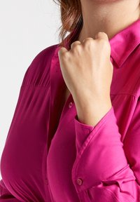 Fuchsia satin blouse with a fitted design, featuring a button-up front, collar, and rolled-up sleeves, showcasing smooth texture and vibrant color.