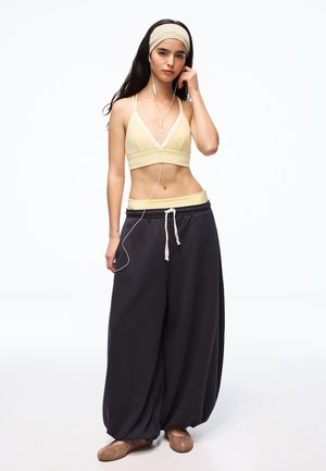 Giovane donna con capelli lunghi e scuri che indossa un reggiseno sportivo giallo, pantaloni della tuta neri e larghi, una fascia per capelli e auricolari, in piedi su uno sfondo bianco.