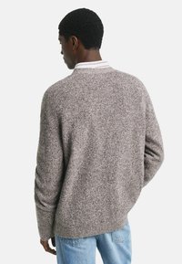 Mann, der einen strukturierten grauen Pullover über einem weißen Kragenhemd und hellblauen Jeans trägt, steht mit dem Rücken zu einem weißen Hintergrund.