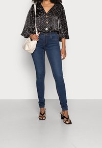 Svart blus med prickar och utsvängda ärmar, knappar, ihop med tajta blå jeans och svarta klacksandaler. Liten krämfärgad handväska.