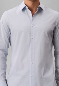 Chemise habillée homme à manches longues bleu clair avec des boutons sur le devant et un petit logo brodé sur la poitrine.