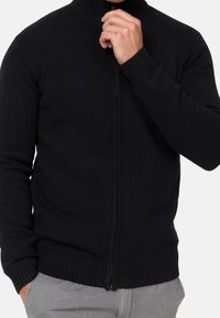 Maglione nero a zip con colletto alto, con polsini e orlo a coste. Presenta una zip marrone e un design di tessuto testurizzato.