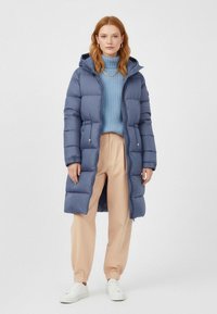 Gevulde lange jas in donkerblauw met capuchon, voorzien van een ritssluiting en zijzakken, gedragen over een lichtblauwe coltrui en beige broek.