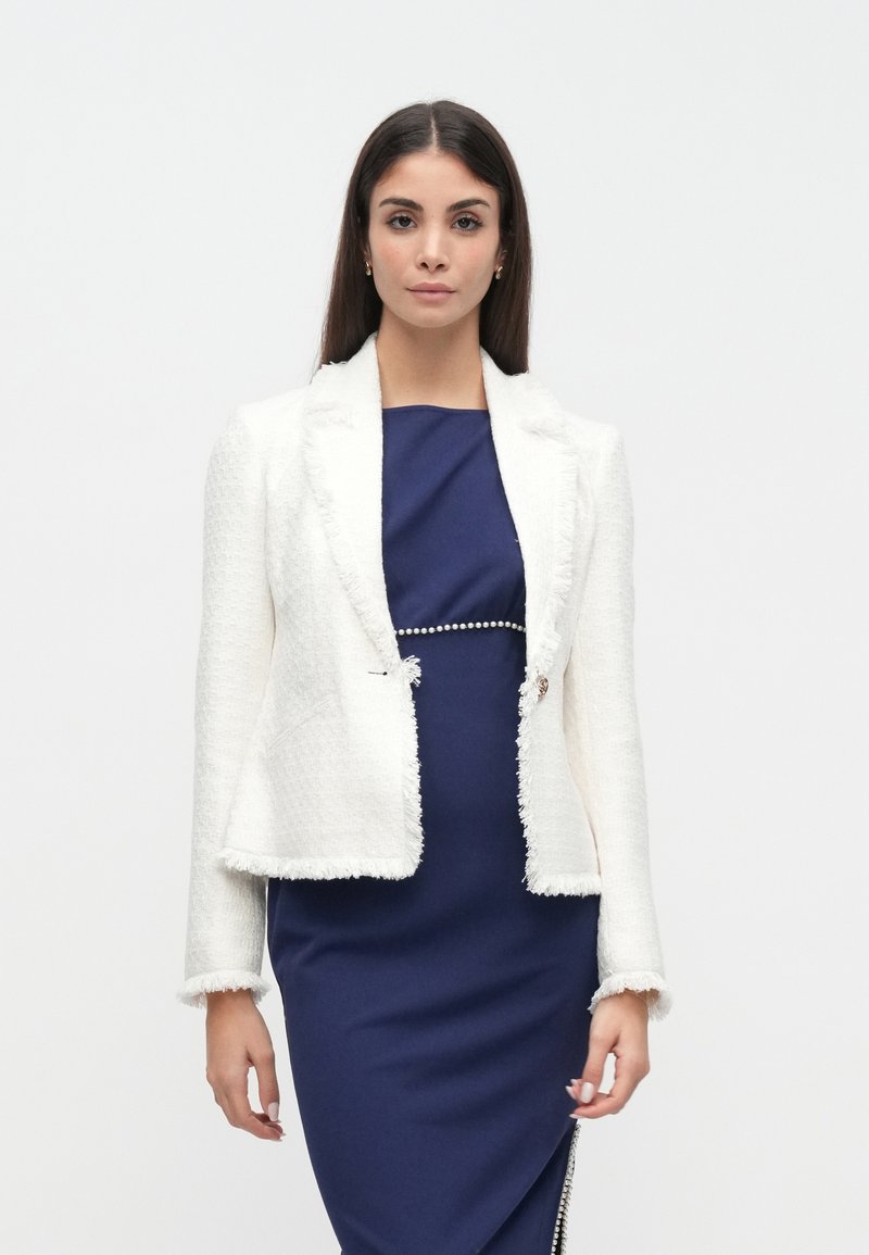 Blazer blanc texturé avec bords effilochés, associé à une robe bleu foncé ajustée ornée d'un accent décoratif en perles à la taille.