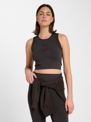 Frau trägt ärmelloses schwarzes Crop-Top und schwarze Leggings mit einem schwarzen Sweatshirt, das um ihre Taille gebunden ist, und steht vor weißem Hintergrund.