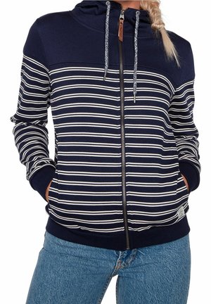 Femme portant un sweat à capuche zippé rayé bleu marine et blanc avec cordons, les mains dans les poches, associé à un jean en denim bleu.