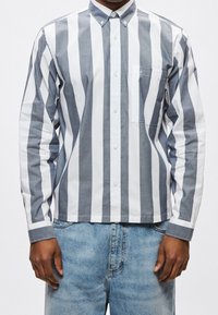 Homme portant une chemise boutonnée à rayures verticales grises et blanches avec une poche poitrine et un jean en denim bleu clair.