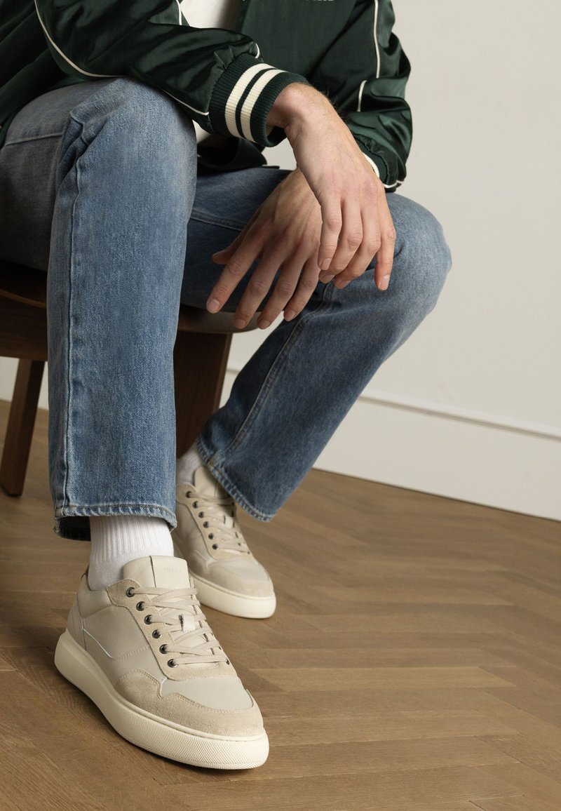 Baskets beiges avec une finition en cuir lisse et en suède, présentant des lacets ronds, une semelle épaisse et des détails de couture subtils, portées avec un jean bleu clair.