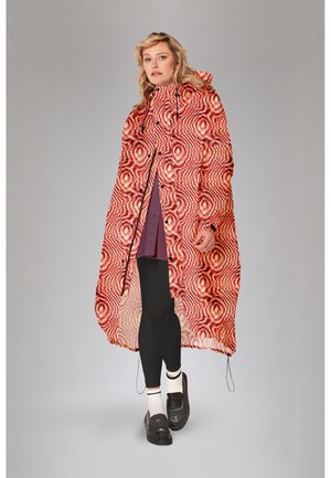 Vrouw die een rode, met patroon bedrukte waterdichte poncho met capuchon draagt, zwarte leggings, witte sokken en zwarte loafers, staande tegen een grijze achtergrond.
