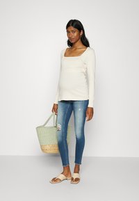 Ljust beige långärmad topp med fyrkantig halsringning, i kombination med slitna blå skinny jeans och en vävd grön väska med en bas av naturlig fiber.