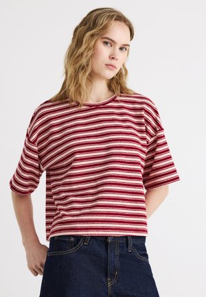 Young woman with wavy blonde hair wearing a red and white striped short-sleeve shirt and dark blue jeans, posing against a white background.