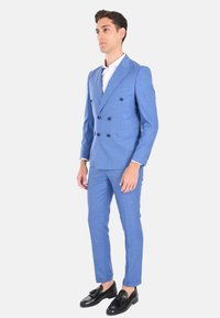 Completo doppiopetto blu chiaro con rever a punta, spalle strutturate e pantaloni slim fit. Indossato con una camicia bianca e mocassini neri.
