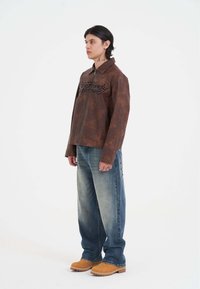 Brun læderjakke med sort tekst, kombineret med lyseblå denimjeans og brune slip-on sko. Jakken har krave og lynlås foran.