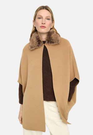 Kobieta w beżowej pelerynie z futrzanym kołnierzem na brązowym swetrze i kremowych spodniach, stojąca na jasnym, jednolitym tle.