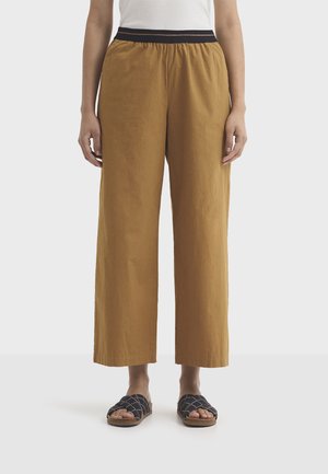 Personne portant un pantalon large ample couleur moutarde avec une taille élastique noire et des sandales noires sans lacets.
