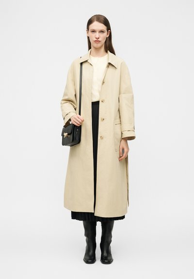 Γυναίκα που στέκεται, ντυμένη με μακρύ μπεζ παλτό, μαύρη πλισέ φούστα, μαύρες μπότες, κρατώντας μια μαύρη τσάντα χιαστί με χρυσή λεπτομέρεια σε λευκό φόντο.
