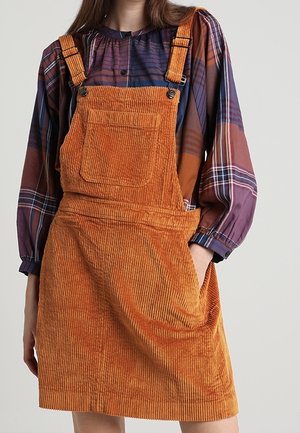 Frau trägt ein orangefarbenes Cord-Overallkleid mit Fronttasche über einer langärmeligen lila karierten Bluse, eine Hand in der Seitentasche.
