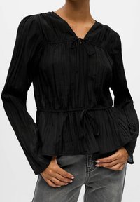 Zwarte geplooide blouse met een V-hals en een strikdetail aan de voorkant, voorzien van lange mouwen en een elastische taille voor extra vorm.